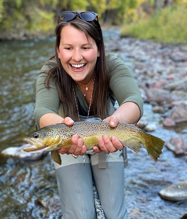 A smiling young girl holds a fish while standing in a creek, enjoying her outdoor Aspen adventure with Wild Willow Outfitters.